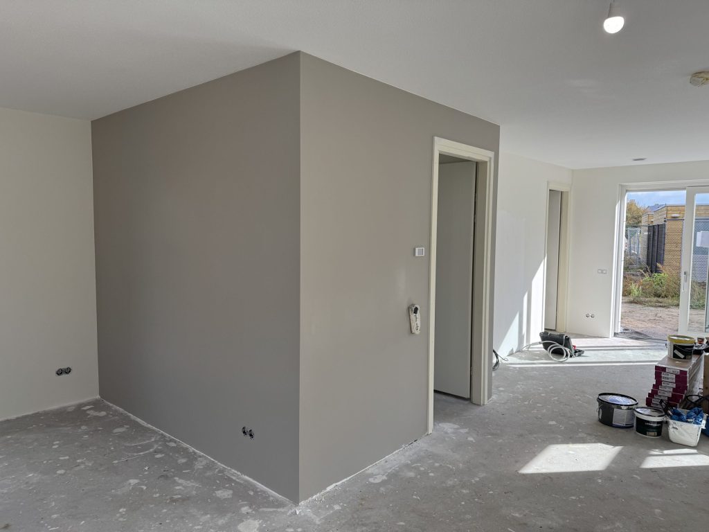 Open woonkamer met een strak gesausde taupe wand rond de meterkast en witte muren naar de tuindeuren