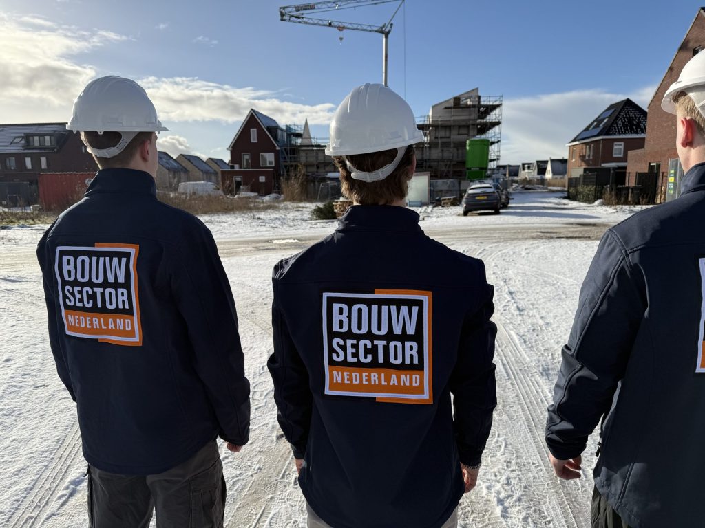 Drie vakmensen van Bouwsector Nederland met witte helm op een besneeuwde straat met huizen in aanbouw